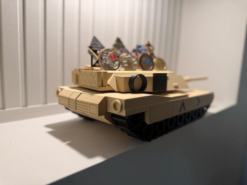 May include: A tan toy tank with black treads and a black circular detail. Several small, decorative items are placed on top of the tank. The tank is sitting on a white shelf.