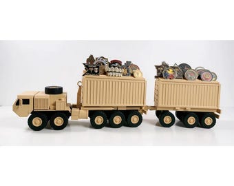 May include: A tan military-style semi-truck with a trailer, featuring a cab and two cargo containers. The tops of the containers are filled with a collection of various circular and shaped metal challenge coins and commemorative pins.