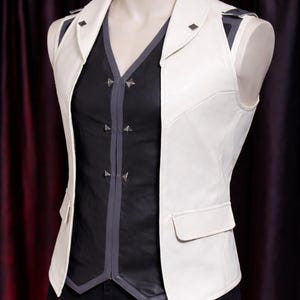 Może przedstawiać: Biała skórzana kamizelka z czarnym i szarym topem. Kamizelka ma kołnierz z klapami, dwie kieszenie i srebrne ćwieki. Czarny top ma dekolt w serek i szare wykończenie. Strój jest prezentowany na manekinie.