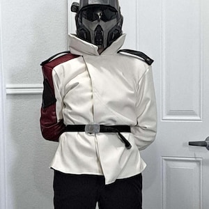 Può includere: Una giacca bianca con dettagli neri e bordeaux, una cintura nera e pantaloni neri. La giacca ha un design asimmetrico unico ed è abbinata a un casco futuristico. L'outfit è un costume cosplay.