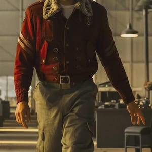 May include: A burgundy bomber jacket with a faux fur collar and multiple button closures. The jacket features two gold stripes on the sleeves and a rectangular pocket. The person is wearing gray cargo pants and a brown belt.