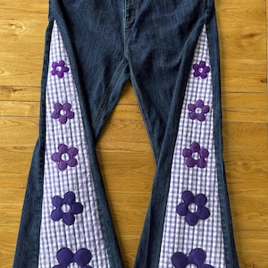 Op de afbeelding: Donkerblauwe denim jeans met uitlopende pijpen. De zijkanten hebben een wit en paars ruitpatroon met grote paarse bloemenapplicaties. Een uniek en stijlvol ontwerp.