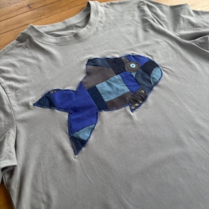 Op de afbeelding: Een grijs t-shirt met een patchwork visontwerp. De vis is gemaakt van verschillende tinten blauw en denim stof, met een kleine blauwe knoop voor het oog. Het t-shirt heeft een ronde hals en korte mouwen.
