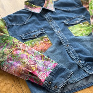 May include: A blue denim shirt with patterned sleeves and collar. The sleeves and collar display a vibrant mix of pink, green, and orange hues. The shirt has two chest pockets and silver snap closures.