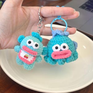 May include: Two handmade crochet keychains. One is a blue bear with a pink bib and cherry design. The other is a blue octopus with a pink mouth. Both have large black eyes and are attached to key rings.