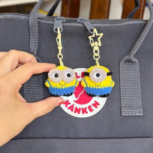 May include: Two handmade crochet keychains featuring yellow and blue characters with round eyes, one with two eyes and one with a single eye. Each keychain is attached to a gold-colored metal clasp, with one featuring a star-shaped attachment.