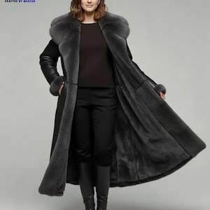 May include: A long, black coat with a gray fur collar and lining. The coat has long sleeves with fur cuffs and a tailored fit. The model is wearing black pants and boots. The logo "CM CRAFTED BY MOAZAM" is in the top left corner.