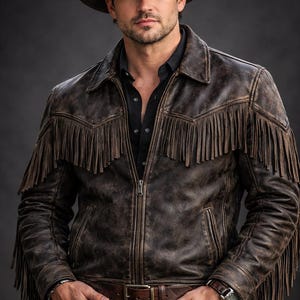 Puede incluir: Chaqueta de cuero marrón con flecos, cinturón a juego y sombrero de vaquero. La chaqueta tiene cierre de cremallera y se lleva sobre una camisa negra y vaqueros azules. Se ve un reloj en la muñeca.