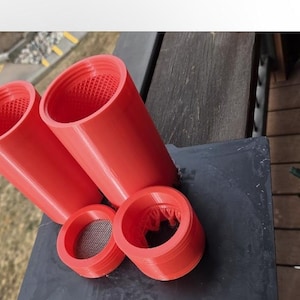 Op de afbeelding: Een verzameling rode, 3D-geprinte cilindrische objecten met gaasinserts. De set bevat twee grote buizen en twee kleinere ringvormige componenten. De objecten zijn gerangschikt op een donker oppervlak, wat wijst op een functioneel of industrieel thema.
