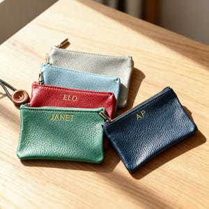 Może przedstawiać: Kolekcja małych, prostokątnych skórzanych portmonetek w różnych kolorach: szarym, jasnoniebieskim, czerwonym, zielonym i granatowym. Każda portmonetka ma złoty zamek błyskawiczny i złote litery z inicjałami lub imieniem. Portmonetki są ułożone na drewnianej powierzchni.