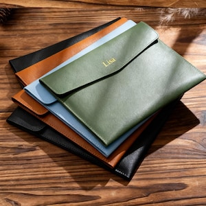 May include: A collection of leather document holders in black, brown, light blue, and green. The green holder is embossed with the name "Lisa" in gold. The items are displayed on a wooden surface.