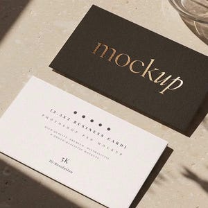 May include: Two business card mockups. One is dark brown with the word "mockup" in gold lettering. The other is white with text that includes "13.3x2 BUSINESS CARD" and "5K Hi-Resolution". The cards are on a light beige surface.