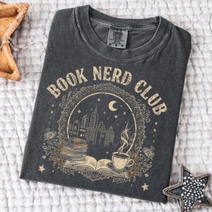 May include: Dark gray t-shirt with the words "BOOK NERD CLUB" in a vintage font. The graphic features a cityscape, books, a coffee cup, and a crescent moon. The shirt is made of a soft, comfortable material.