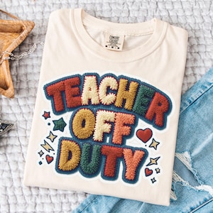 Peut inclure: T-shirt crème avec l'inscription "TEACHER OFF DUTY" dans un design coloré et texturé. Le motif comprend des accents rouges, oranges, jaunes, verts et bleus, ainsi que des graphiques d'étoiles et de cœurs. Le t-shirt est plié sur une surface blanche.