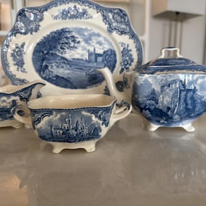 Puede incluir: Juego de té de cerámica azul y blanca con un diseño de paisaje. El juego incluye un plato, una azucarera, una jarra de crema y dos tazas de té. Las piezas tienen un borde festoneado y están sobre una superficie reflectante.