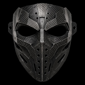 Könnte beinhalten: Eine taktische Vollgesichtsmaske mit dunkelgrauem Carbonfasermuster und einer mehrteiligen, kantigen Konstruktion. Die Maske verfügt über zwei große Augenöffnungen, einen zentralen Nasensteg und einen unteren Bereich mit vertikalen Reihen kreisförmiger Belüftungslöcher.