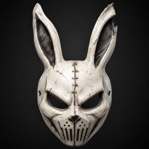 May include: A white rabbit mask with a weathered, cracked finish and dark shading. The mask features long ears, one with a chipped top, a stitched seam down the center, large eye openings, a small nose, and a vertical slatted mouth area.
