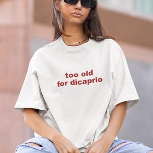 Könnte beinhalten: Weißes T-Shirt mit dem roten Text "too old for dicaprio". Es wird mit hellblauen zerrissenen Jeans und weißen Turnschuhen getragen. Die Person trägt eine schwarze Sonnenbrille und eine goldene Halskette.