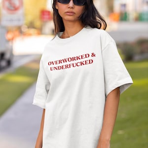 May include: White t-shirt with the text "OVERWORKED & UNDERFUCKED" in red. The shirt is worn by a person wearing black sunglasses. The background is blurred, suggesting an outdoor setting.