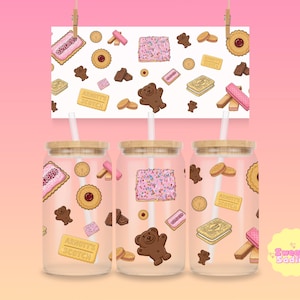 May include: Three glass tumblers with bamboo lids and straws, featuring a repeating pattern of illustrated cookies, including pink frosted biscuits, bear-shaped cookies, and shortbread. A matching print hangs above. The tumblers have a pink background.