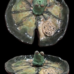 May include: Handmade ceramic frog dish with a lily pad design. The frog is green with large eyes, sitting on a green and brown lily pad. The dish has a glossy finish and is suitable for holding small items.