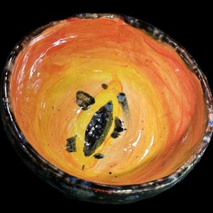 Peut inclure: Un bol en céramique fait à la main avec un intérieur orange et jaune vif, orné de taches sombres et d'un motif central noir et blanc. Le bord du bol est bleu et noir foncé et texturé. Le fond est noir uni.