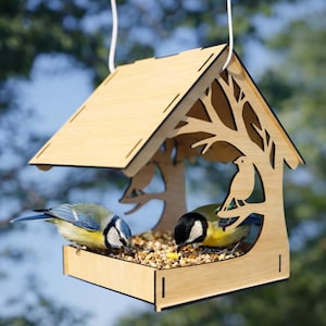 Puede incluir: Un comedero para pájaros de madera con un diseño recortado en forma de árbol, que alberga dos pájaros pequeños. El comedero está suspendido por un cordón blanco y lleno de alpiste. Las aves tienen plumaje azul, amarillo y negro.