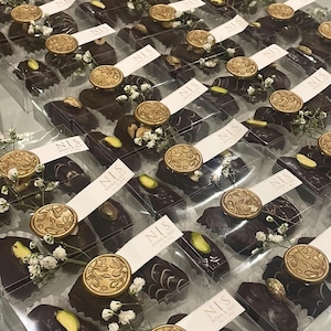 Könnte beinhalten: Draufsicht auf zahlreiche transparente Schachteln mit dunklen Schokoladenkonfekt. Jede Schachtel ist mit einem goldenen Siegel, kleinen weißen Blumen und einem Etikett mit der Aufschrift "NIS NORA & SEIF" verziert. Einige Pralinen haben Nüsse.