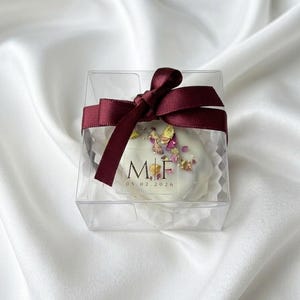Könnte beinhalten: Eine transparente, quadratische Box, gebunden mit einem weinroten Band, enthält eine weiße, herzförmige Leckerei. Die Leckerei ist mit getrockneten Blumen und den Initialen "M.F" und dem Datum "05.02.2026" verziert. Die Box liegt auf einer weißen, seidigen Oberfläche.