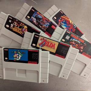 Puede incluir: Una colección de cartuchos de juegos de Super Nintendo. Los cartuchos grises tienen etiquetas coloridas con títulos de juegos como "Mario World", "The Legend of Zelda" y "Donkey Kong Country". Las etiquetas muestran varios personajes y arte del juego.