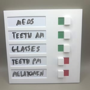 May include: A white rectangular task tracker board with five horizontal slots labeled in black marker: MEDS, TEETH AM, GLASSES, TEETH PM, and MELATONIN. Each slot has a sliding white toggle switch that reveals either a green or red square indicator.
