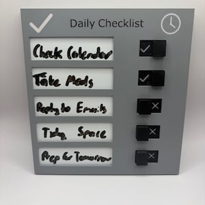 May include: A gray daily checklist board with white inserts featuring handwritten tasks including Check Calendar, Take Meds, Reply to Emails, Tidy Space, and Prep for Tomorrow. The board includes five black sliding toggles to mark items as complete.
