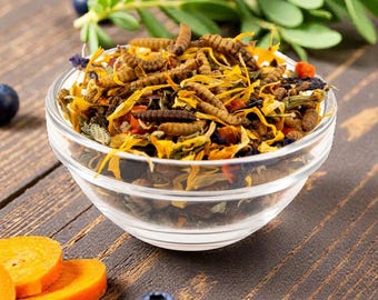 May include: A clear glass bowl filled with a mix of dried insects, yellow flower petals, and other colorful ingredients. Sliced carrots and fresh blueberries are scattered around the bowl on a wooden surface. Green sprigs are in the background.