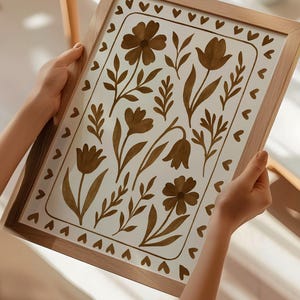 May include: A framed floral art print featuring brown flowers and leaves on a white background. The artwork is enclosed in a light brown wooden frame with a border of small brown hearts. The print is held by two hands.