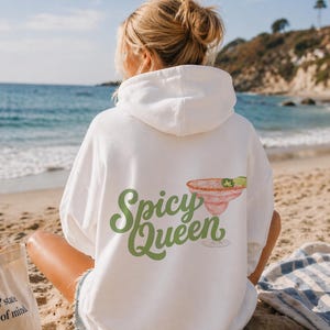 Puede incluir: Sudadera con capucha blanca con la frase "Spicy Queen" en verde y una ilustración de margarita. La sudadera se lleva en una playa con el mar de fondo.