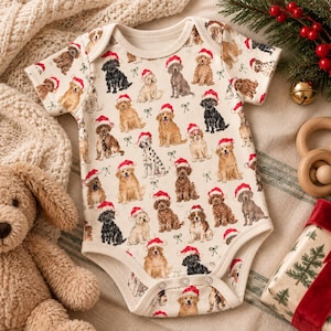 May include: A short-sleeved baby bodysuit in an off-white color featuring an all-over print of various dog breeds wearing red and white Santa hats, interspersed with small green bows. The garment has a round neckline and snap closures at the bottom.