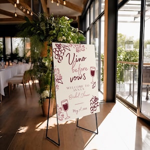 May include: A white rectangular sign on a black metal stand featuring the text Vino before vows, Welcome to Anna's Bridal Shower, and May 31st, 2026. The sign is decorated with maroon illustrations of wine glasses and clusters of grapes.