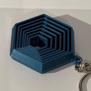May include: A teal blue, multi-layered, hexagonal keychain charm. The charm has a series of nested hexagonal shapes, creating a 3D effect. A silver-colored keyring is attached to the charm. The keychain is on a white surface.