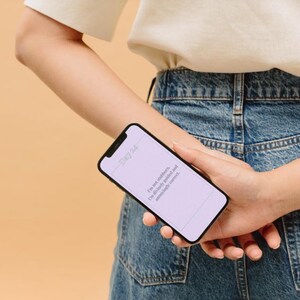 Op de afbeelding: Een zwarte smartphone met een lichtpaars scherm met tekst wordt achter de rug gehouden. De telefoon wordt vastgehouden tegen een achtergrond van een persoon die een wit shirt en een blauwe spijkerbroek draagt. De tekst op het scherm luidt: "Ik ben niet koppig. Ik..."