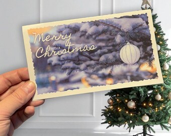 May include: A festive Christmas card with the words "Merry Christmas" in cursive. The card shows a snowy evergreen branch with a white bauble. A decorated Christmas tree with lights and baubles is visible in the background.