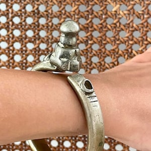 Può includere: Un bracciale a polsino color argento con un design aperto. Il bracciale presenta una figura dettagliata sulla parte superiore e segni incisi vicino all'apertura. Il gioiello è indossato su un polso, mostrando il suo design e la sua maestria.