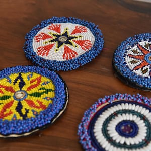 May include: Four round beaded coasters with intricate designs. Each coaster features a central pattern in various colors, including red, white, yellow, and blue, surrounded by a border of blue beads. The coasters are displayed on a wooden surface.