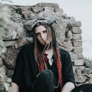 May include: A person wearing a black outfit, seated against a stone wall. The outfit includes a long-sleeved top, pants, and boots. Accessories include a horned headpiece, jewelry, and wrist cuffs. Red streaks are in the person's long hair. The text "DANCING TRIBE" is at the bottom.