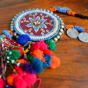 Puede incluir: Un collar de cuentas de colores con un gran colgante redondo que presenta un diseño de estrella. El colgante está decorado con cuentas, conchas y detalles plateados. El collar tiene una larga borla de hilo de colores.