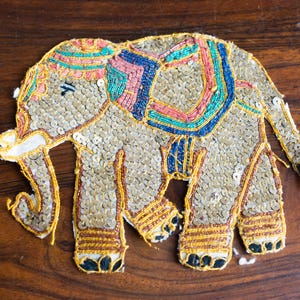 May include: A decorative elephant applique, covered in gold sequins, with colorful beaded accents in pink, blue, and green. The elephant has a detailed trunk, legs, and ears, and is set against a dark wood background.