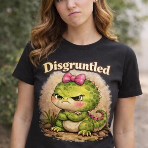 May include: Black t-shirt featuring a cartoon dinosaur with an angry expression and a pink bow. The word "Disgruntled" is printed above the dinosaur in gold lettering. The dinosaur is green with pink accents.