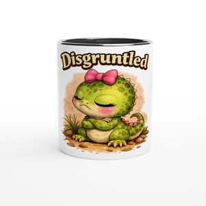 May include: White ceramic mug with a black interior rim. The mug features a cartoon illustration of a green dinosaur with a pink bow, crossed arms, and the word "Disgruntled" in brown lettering.
