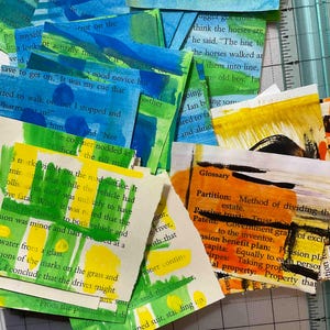 May include: A collection of rectangular paper scraps featuring printed book text overlaid with abstract brushstrokes in shades of blue, green, yellow, and orange. The pieces are scattered across a cutting mat with visible inch measurement markings.