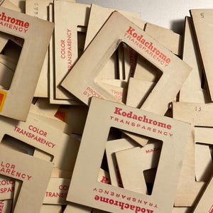 May include: A collection of square cardboard Kodachrome transparency slide mounts stacked in a pile. Each off-white mount features red printed text reading Kodachrome Transparency and Color Transparency, with some showing the yellow Kodak logo.