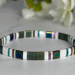 May include: A beaded bracelet featuring square, iridescent beads in shades of green, white, teal, and purple. The bracelet is strung together, creating a flexible and stylish accessory.